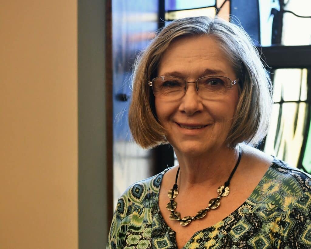 A woman in glasses standing in front of a stained glass window.