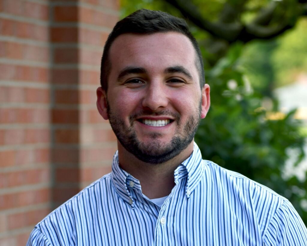 A man in a striped shirt smiling in front of a brick wall.