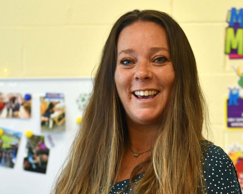 A woman in a polka dot shirt smiles in front of a bulletin board.