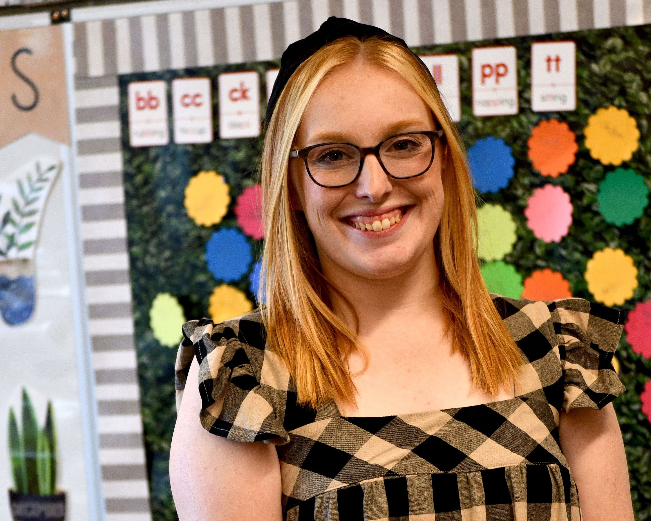A girl in glasses smiles in front of a bulletin board.