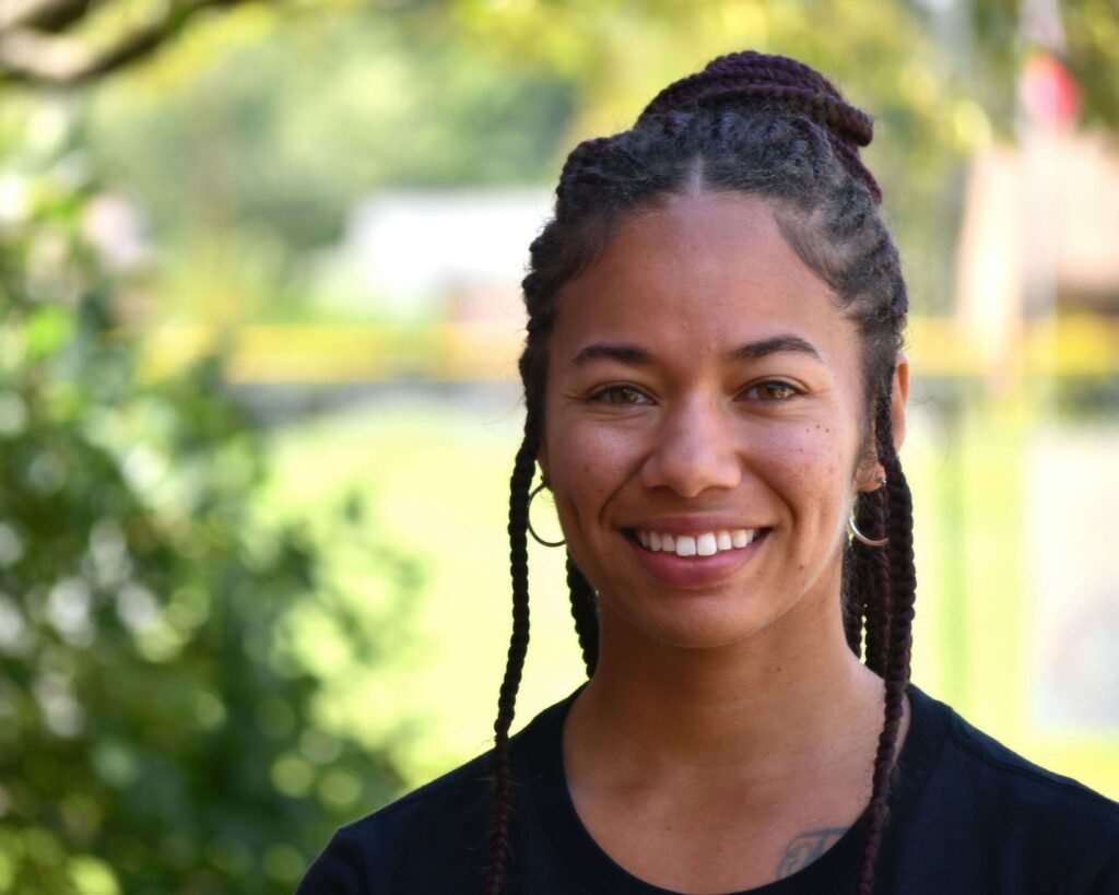 A woman with braids smiling in front of a tree.