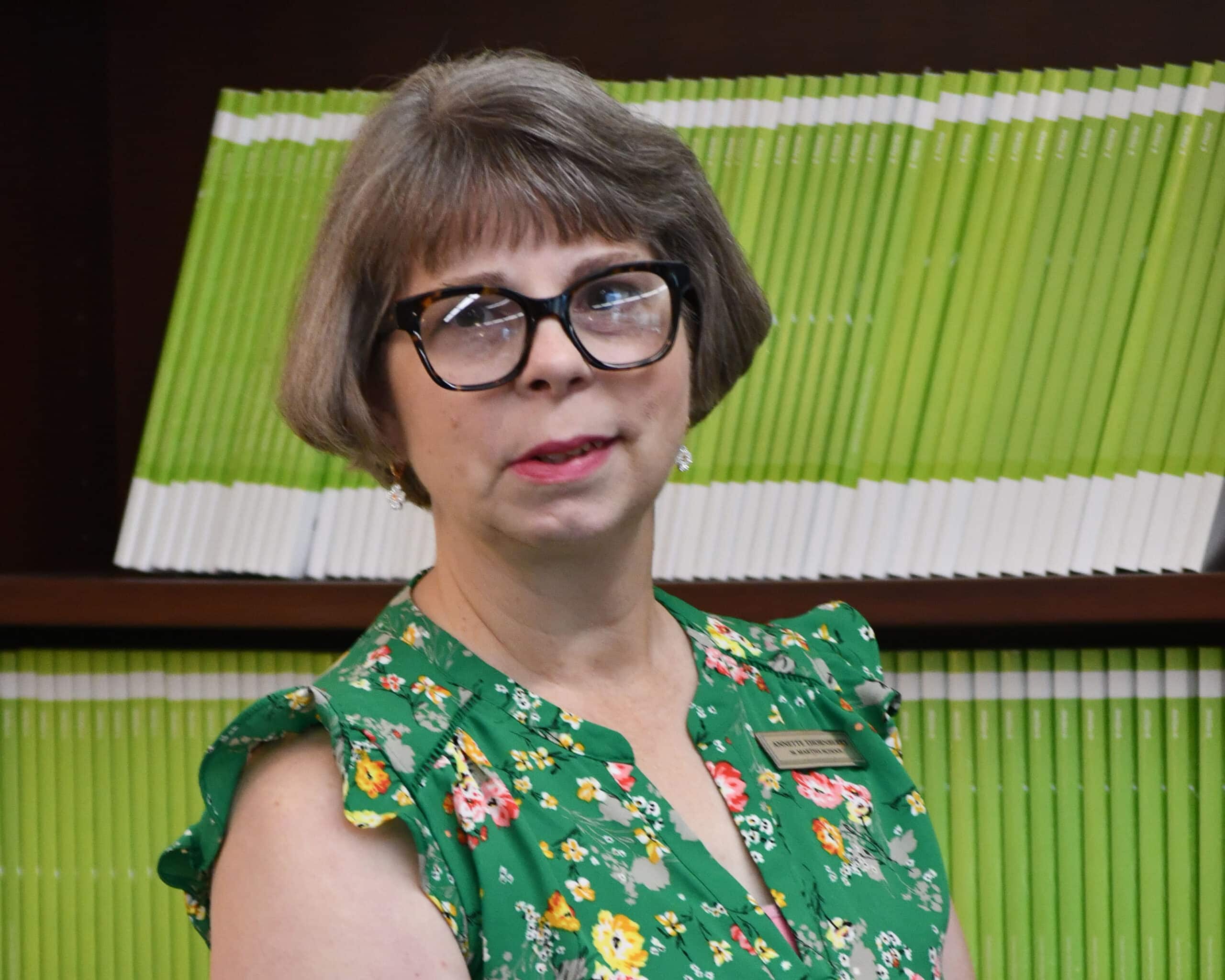 A woman in glasses standing in front of a green book shelf.