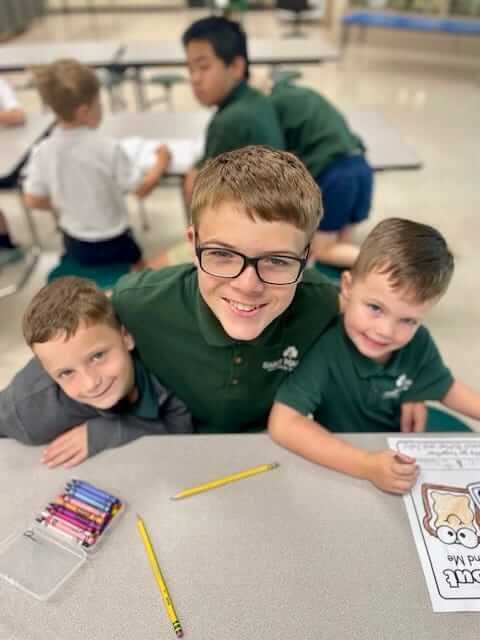 Three young boys in green school uniforms sit at a table coloring with crayons and pencils. Other children work in the background.