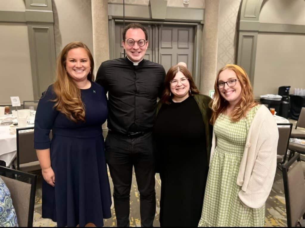 Four people posing for a photo indoors, including a man in a black clerical shirt and three women.