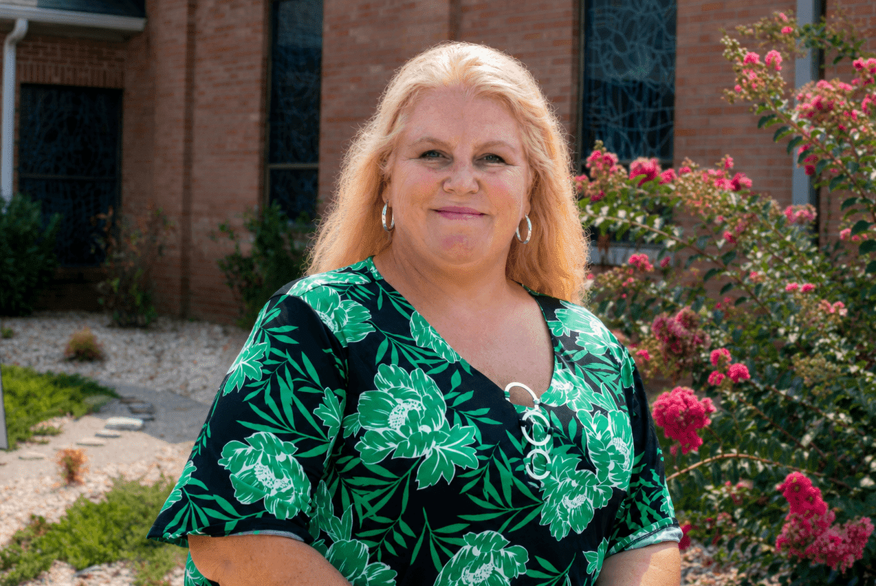 A woman with long blonde hair wearing a green and black floral print top stands outside a brick building next to a blooming crape myrtle bush.