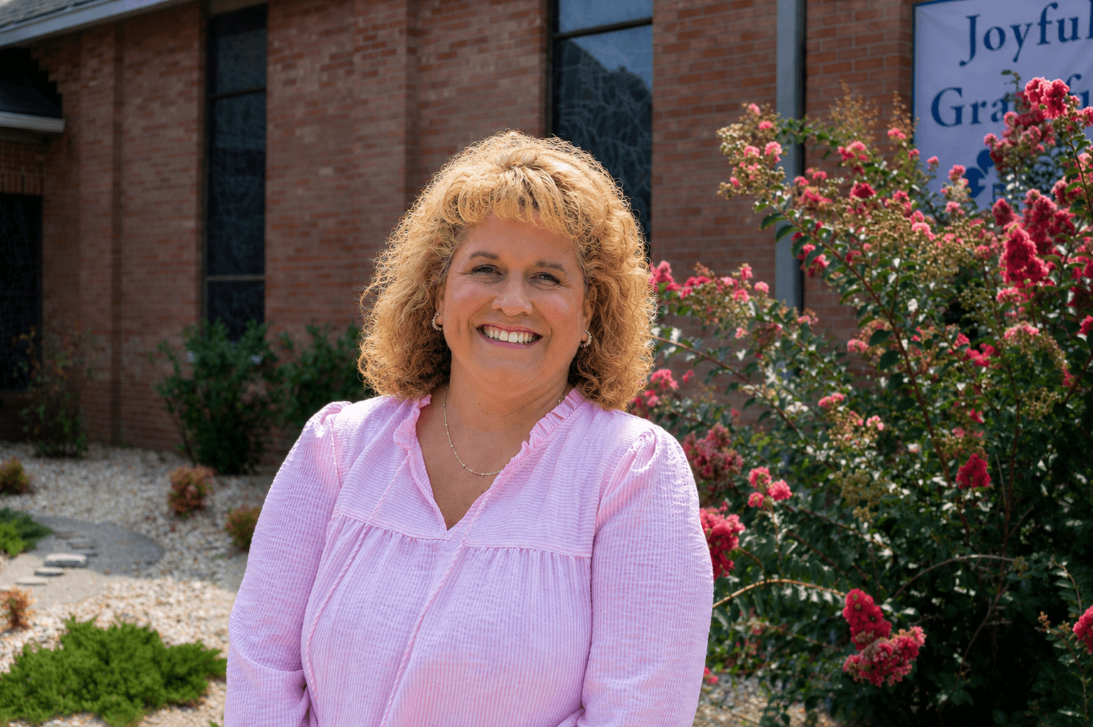 A smiling woman with blonde hair wearing a light pink shirt, standing in front of a brick building and a flowering bush.