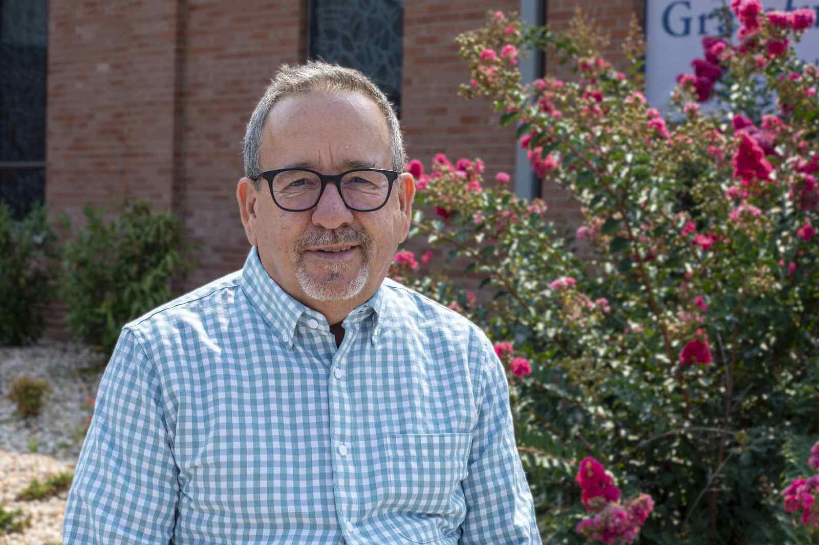 Middle-aged man with glasses and a goatee, wearing a light-colored checkered button-down shirt, standing outdoors in front of a blooming crape myrtle tree.