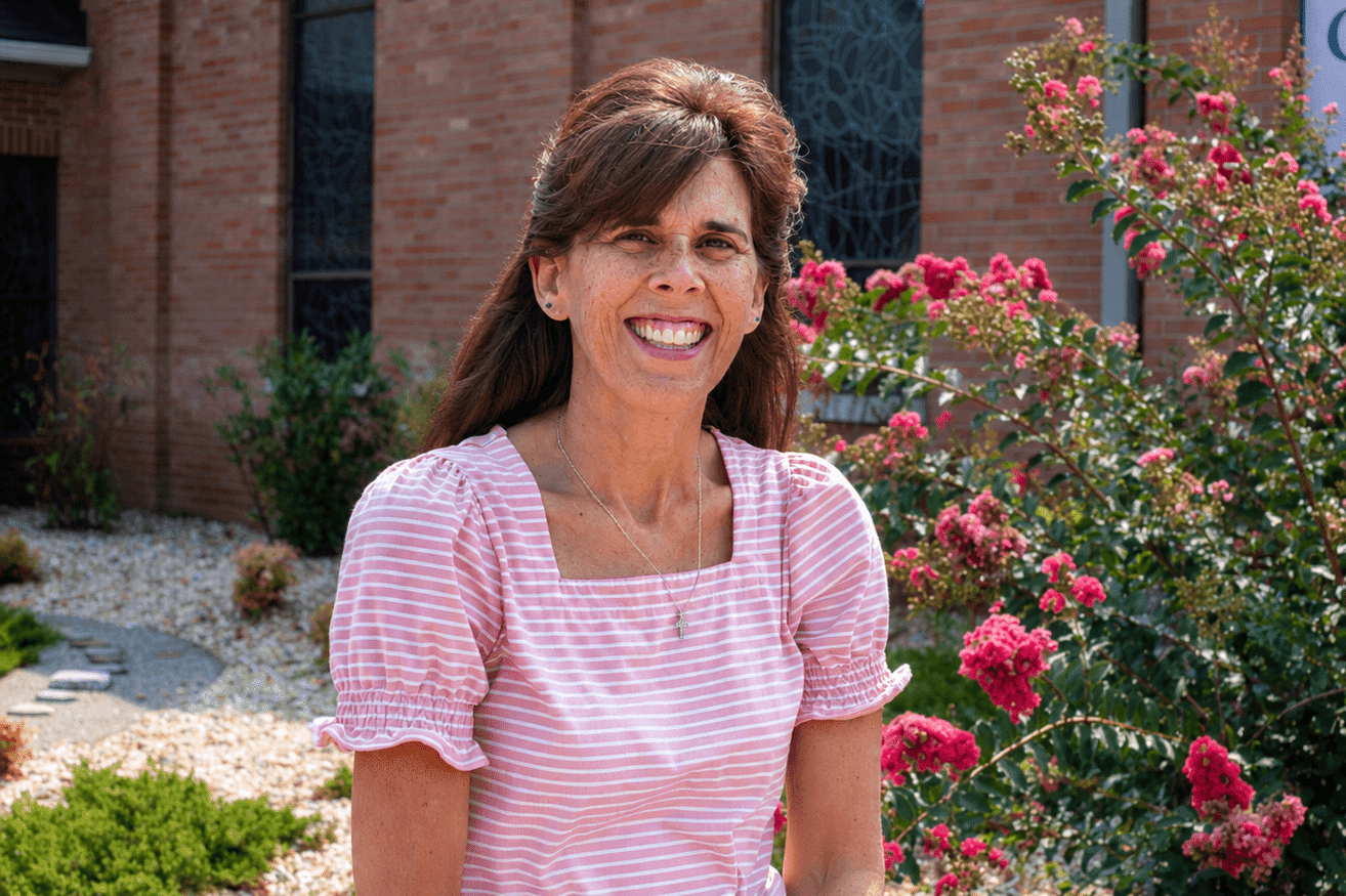 A smiling woman in a pink striped shirt standing in front of a brick building and a blooming crape myrtle tree.