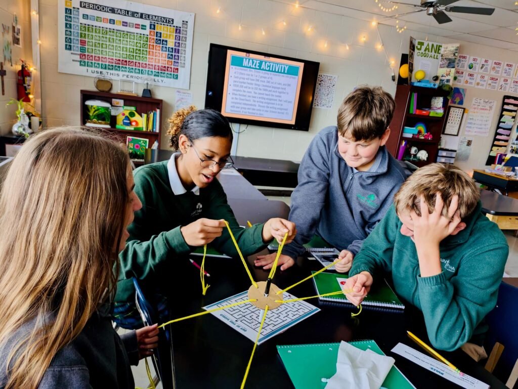 Four students collaborate at a table to navigate a central wooden hub using attached strings to move a pen through a maze.