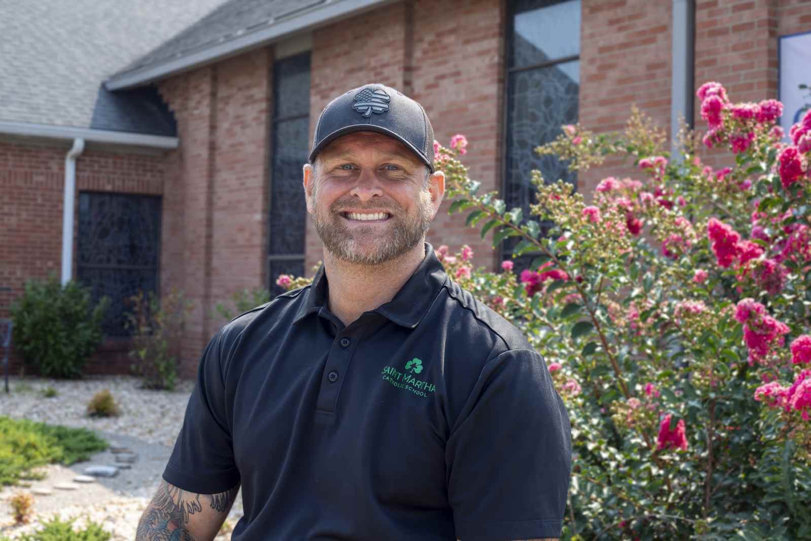 A man wearing a black Saint Martha Catholic School polo shirt and a baseball cap stands outside a brick building near a flowering bush.