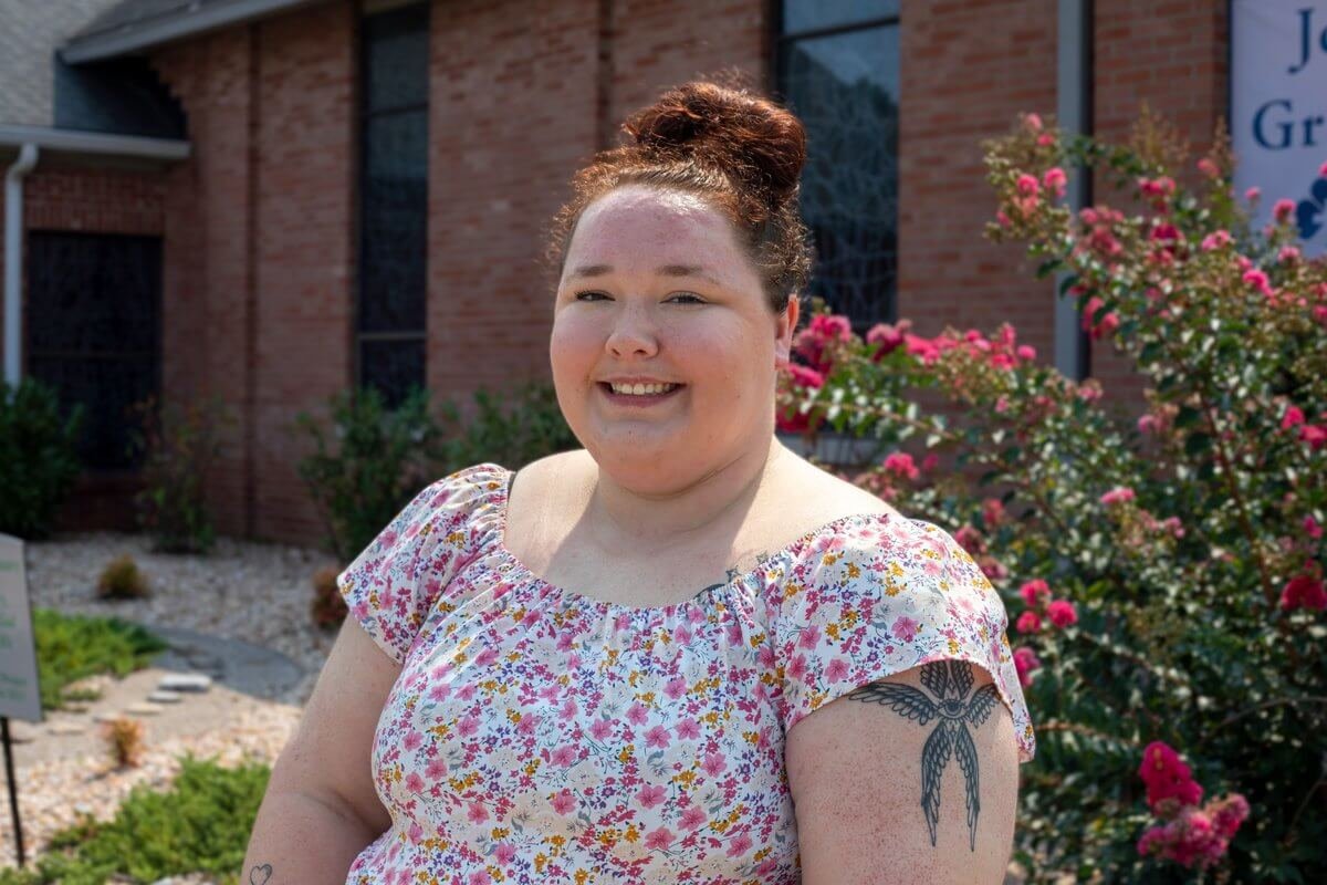 A smiling woman with her hair in a bun wearing a floral print top stands outdoors in front of a brick building.