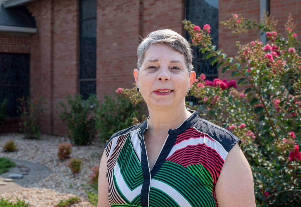A woman with short hair wearing a patterned sleeveless top stands in front of a brick building and a flowering bush.