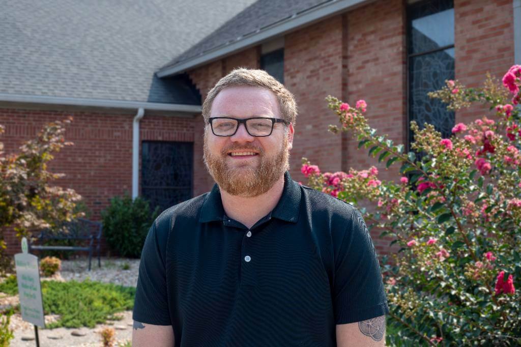 A smiling man with a red beard and glasses wearing a black polo shirt, standing outside a brick building with pink flowering bushes.